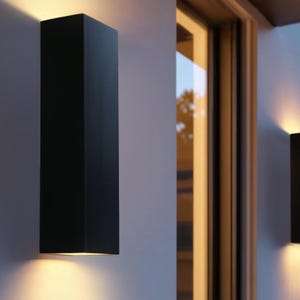 May include: A pair of black, rectangular outdoor wall sconces. Each sconce has a downward-facing light source, casting a warm glow on the wall below. The sconces are mounted on a light-colored wall near a window.
