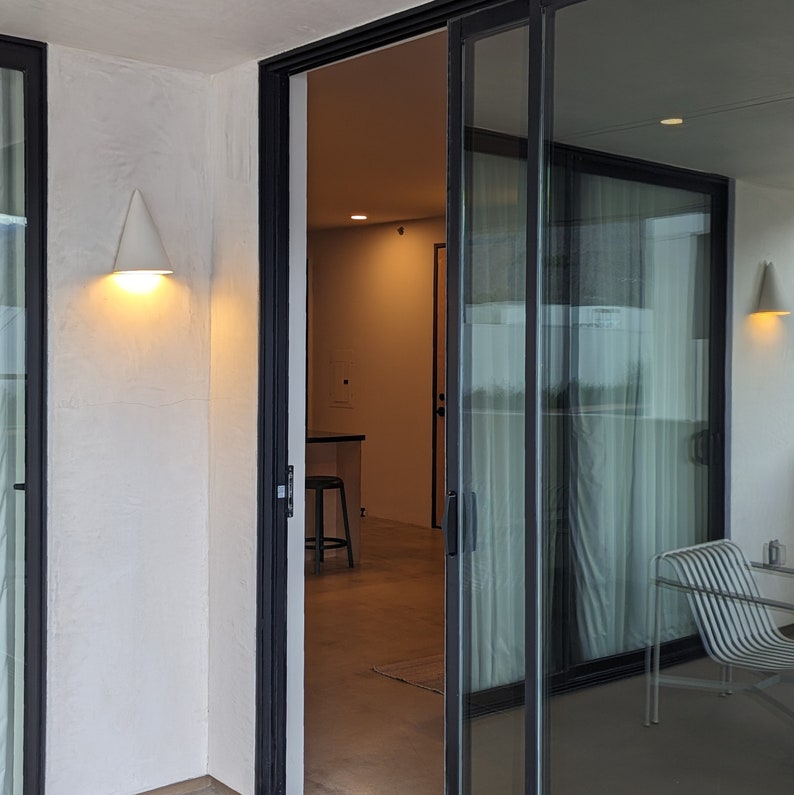 May include: A white wall with a white light fixture on the left side of the image. A black door with a glass panel leads to a room with a white wall and a black door. The glass panel is reflecting the room and a white chair.