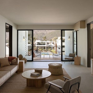 May include: A living room with a large window overlooking a pool and patio area. The room is furnished with a couch, two chairs, a coffee table, and a woven ottoman. The walls are white and the floor is a light gray concrete.