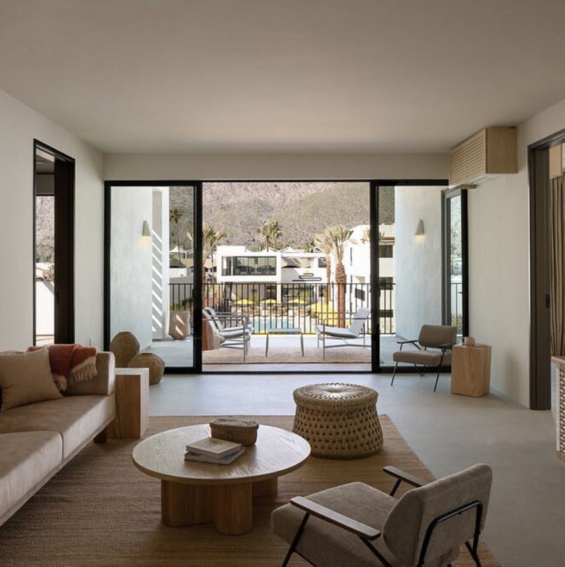 May include: A living room with a large window overlooking a pool and patio area. The room is furnished with a couch, two chairs, a coffee table, and a woven ottoman. The walls are white and the floor is a light gray concrete.