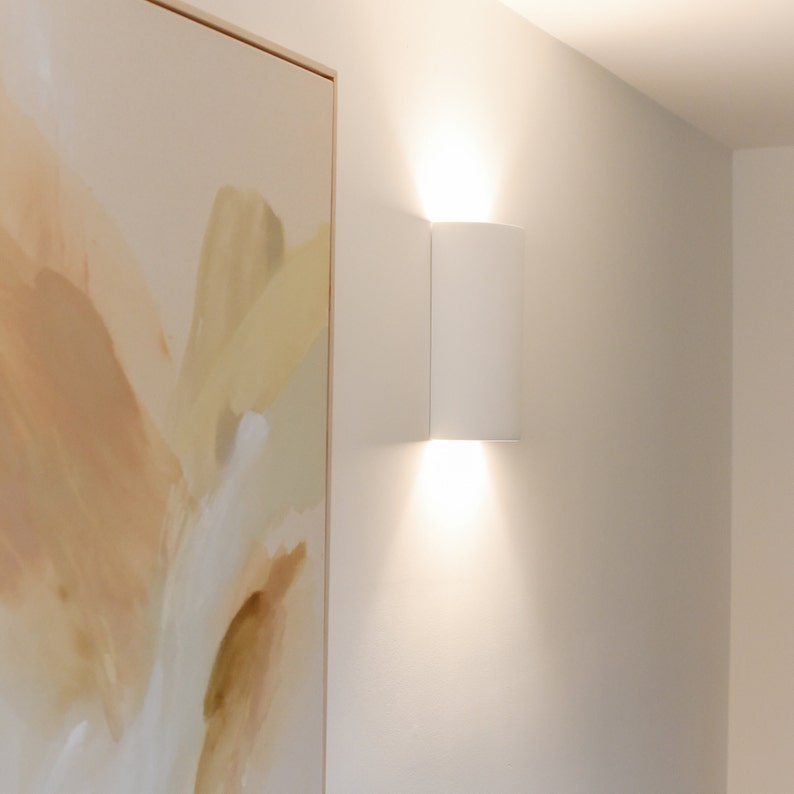 K&ouml;nnte beinhalten: Eine wei&szlig;e, rechteckige Wandleuchte mit einem sanften, wei&szlig;en Licht. Die Leuchte ist neben einem gerahmten abstrakten Gem&auml;lde in Beige- und Braunt&ouml;nen angebracht.