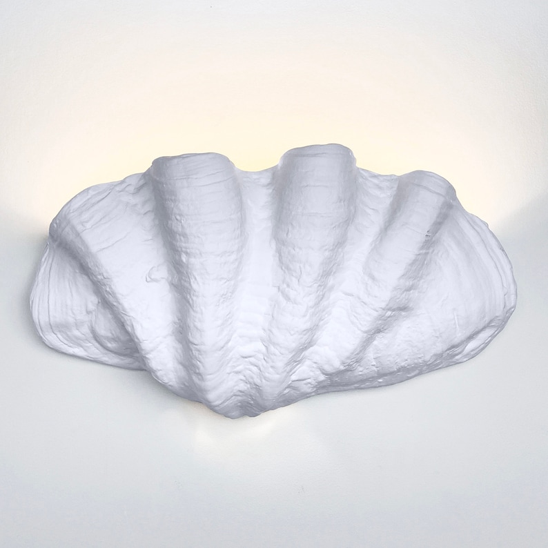 May include: A white ceramic wall sconce shaped like a large seashell. The sconce has a textured surface and is lit from within.