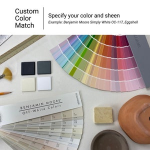 May include: A color chart with a variety of paint colors, including off-white, beige, and gray. The chart is labeled "Benjamin Moore Off-White Colors". There are also paint chips, a paint brush, a pencil, and a small terracotta pot.