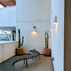 May include: A white wall with two white cone-shaped lights and a fire alarm. There are two potted plants, a bench, and a white wall in the background.