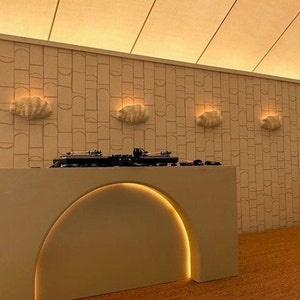 May include: A white DJ booth with an arched opening and a yellow light strip. The booth is in front of a white wall with a geometric pattern and four white shell-shaped wall sconces.