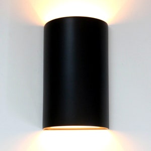K&ouml;nnte beinhalten: Eine schwarze zylindrische Wandleuchte mit einem warmwei&szlig;en Licht, das von unten ausstrahlt.