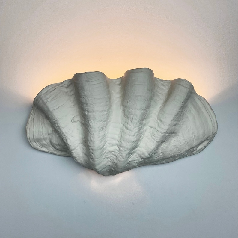 May include: A white ceramic wall sconce shaped like a large seashell. The sconce has a textured surface and is lit from within.
