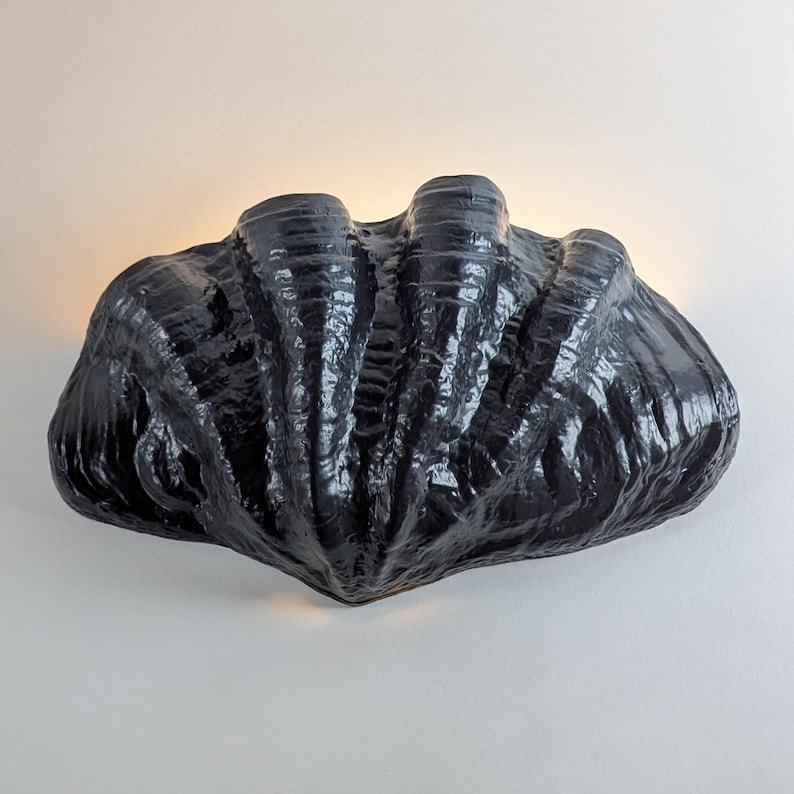 May include: A black ceramic wall sconce shaped like a large clam shell. The sconce has a textured surface and is lit from within.