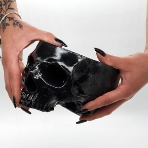 May include: A black skull-shaped planter is held in two hands. The skull is glossy and has a detailed design.
