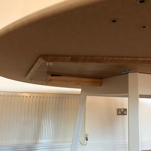 May include: The underside of a round, wooden table with a white metal base. The table top is made of light brown wood and has a slightly rounded edge. The base is made of white metal and has a simple, modern design.