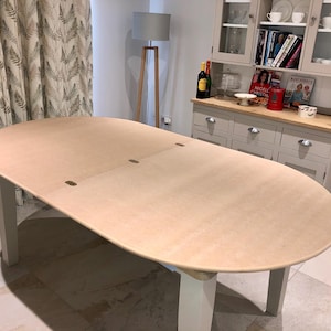 May include: An oval dining table with a light wood surface and white legs. The table has a smooth, unfinished appearance and is set in a well-lit room. The table's dimensions are approximately 183 cm long and 102 cm wide.
