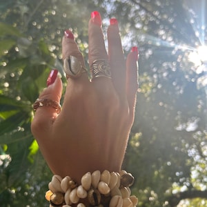 May include: A hand adorned with rings and bracelets. The rings include a gold band with a shell, and a gold ring with a sun design. The bracelets are made of white seashells and gold accents. The nails are painted a bright red.
