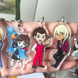 May include: Five colorful cartoon character keychains with clear acrylic backgrounds. The characters are a blonde man in a blue suit, a girl with brown hair wearing a blue cape, a man with brown hair in a red suit, a blonde man in a red jacket, and a girl with brown hair wearing a white lab coat.
