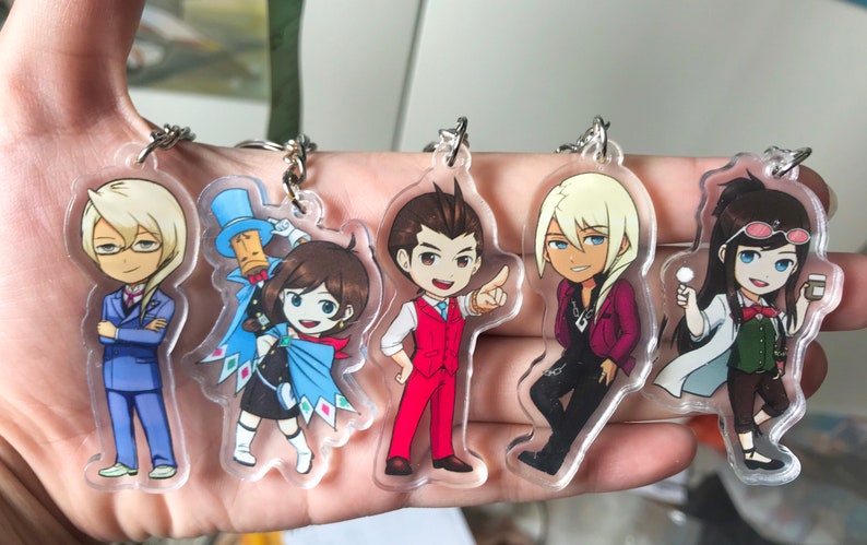May include: Five colorful cartoon character keychains with clear acrylic backgrounds. The characters are a blonde man in a blue suit, a girl with brown hair wearing a blue cape, a man with brown hair in a red suit, a blonde man in a red jacket, and a girl with brown hair wearing a white lab coat.