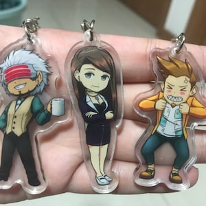 May include: Three clear acrylic keychains with cartoon character illustrations. The keychain on the left features a man with white hair, a red headband, and a cup of coffee. The keychain in the middle features a woman with long brown hair, wearing a black dress and a necklace. The keychain on the right features a man with brown hair, wearing a yellow jacket and a white shirt.