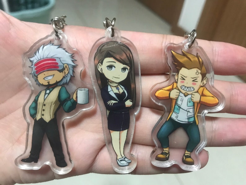 May include: Three clear acrylic keychains with cartoon character illustrations. The keychain on the left features a man with white hair, a red headband, and a cup of coffee. The keychain in the middle features a woman with long brown hair, wearing a black dress and a necklace. The keychain on the right features a man with brown hair, wearing a yellow jacket and a white shirt.