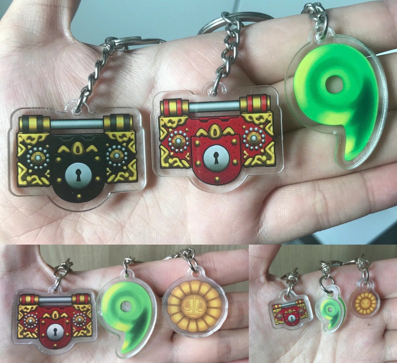 May include: Three sets of keychains featuring a green swirl, a gold and red chest with a keyhole, and a gold coin with a scale design. The keychains are made of clear acrylic and have a silver metal ring.