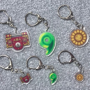 May include: Six keychains with different charms. Two charms are red and gold with a keyhole, one is green and shaped like a number nine, and three are gold with a sun and scales design.
