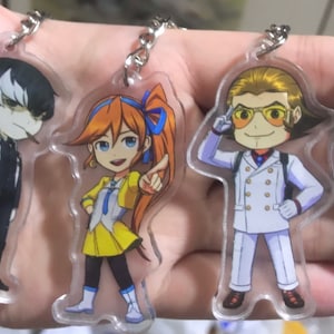 May include: Four colorful anime character keychains with a clear acrylic background. The characters are a girl with orange hair and a yellow dress, a boy with blonde hair wearing a white suit, a boy with black hair wearing a blue suit, and a boy with black hair wearing a black suit with a bird on his shoulder.