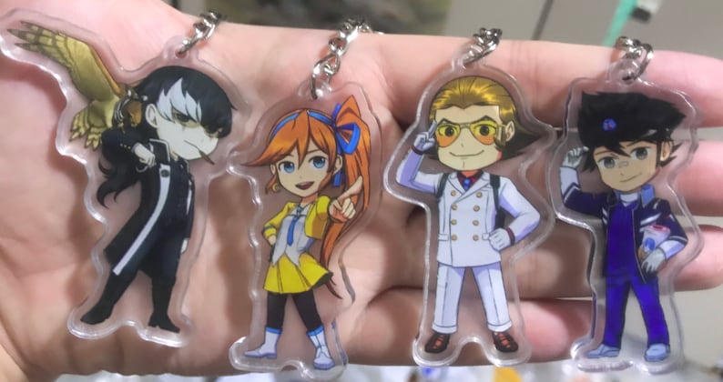 May include: Four colorful anime character keychains with a clear acrylic background. The characters are a girl with orange hair and a yellow dress, a boy with blonde hair wearing a white suit, a boy with black hair wearing a blue suit, and a boy with black hair wearing a black suit with a bird on his shoulder.