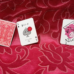 Könnte beinhalten: Drei Spielkarten auf einer roten, gemusterten Oberfläche. Eine Karte zeigt ein rotes Design, eine andere ein Pik-Ass mit Blutspritzern und die dritte ein Cartoon-Gesicht mit dem Namen "Nick".