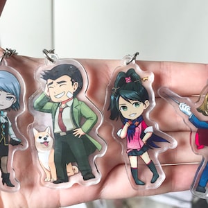 May include: Four anime-style character keychains. The characters are a blue-haired girl with a black dress and a white collar, a brown-haired man with a green coat and a red tie, a green-haired girl with a pink dress and a blue cape, and a brown-haired boy with a blue suit and a red cape.