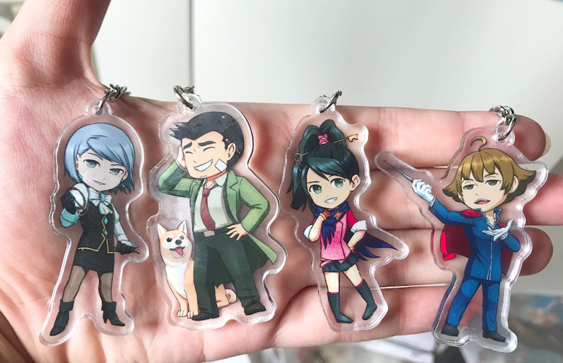May include: Four anime-style character keychains. The characters are a blue-haired girl with a black dress and a white collar, a brown-haired man with a green coat and a red tie, a green-haired girl with a pink dress and a blue cape, and a brown-haired boy with a blue suit and a red cape.