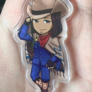May include: A cartoon character keychain with a cowboy hat, blue jacket, and red bandana. The character is wearing brown boots and a tan poncho with blue and white patterns.