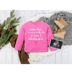 May include: Pink long-sleeved shirt with white text that reads "Lieber Papa ich wünsche Dir alles Gute zu Deinem 1. Geburtstag mit mir" and a red heart.