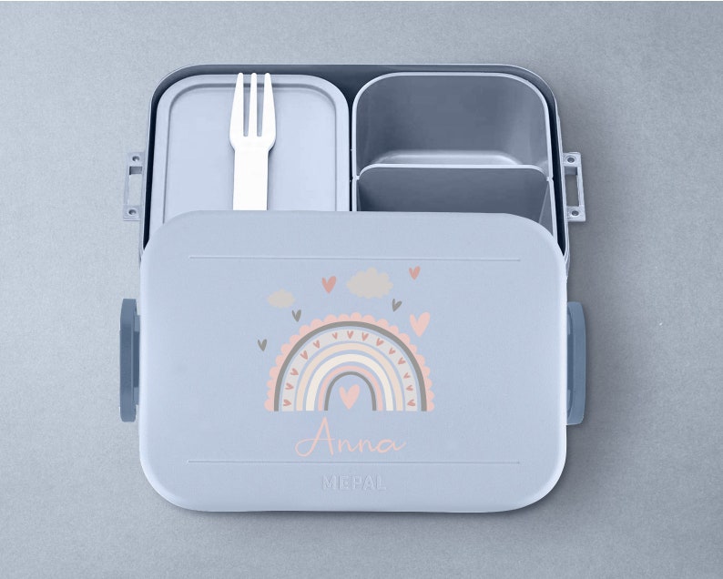 May include: A light blue lunchbox with a rainbow and cloud design. The lunchbox has a removable fork and a compartment for snacks. The lunchbox is personalized with the name "Anna".