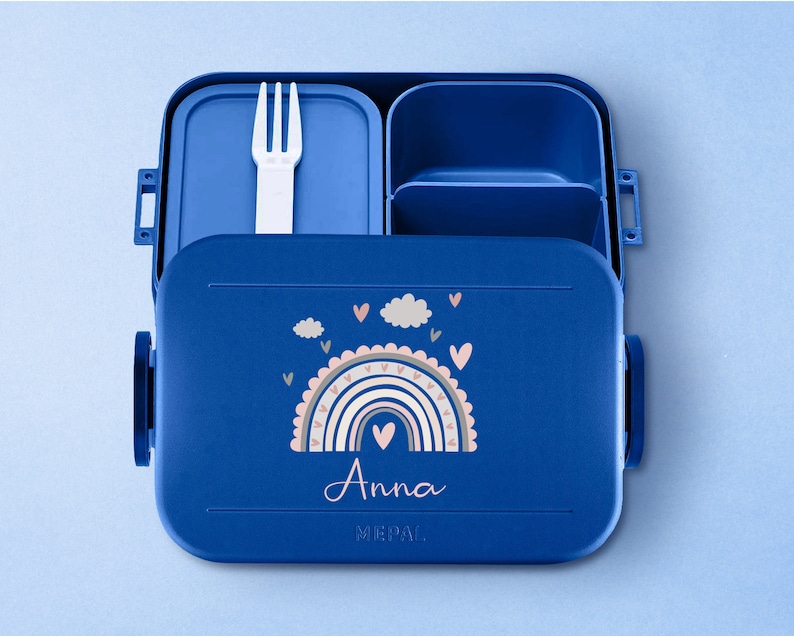 May include: Blue lunchbox with a rainbow and cloud design. The lunchbox has a removable fork and a compartment for snacks. The lunchbox is personalized with the name "Anna".