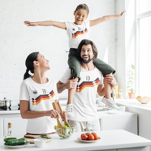 Könnte beinhalten: Eine dreiköpfige Familie trägt weiße T-Shirts mit der deutschen Flagge und aufgedruckten Nummern. Der Vater trägt die Tochter auf seinen Schultern. Die Mutter bereitet in der Küche einen Salat zu.