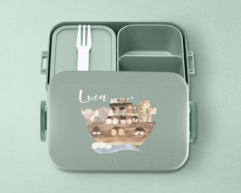 Puede incluir: Una fiambrera verde con tapa extra&iacute;ble y un compartimento separado para cubiertos. La fiambrera tiene una ilustraci&oacute;n colorida de animales en un barco y el nombre "Luca" impreso en la parte delantera. La fiambrera est&aacute; hecha por Mepal.