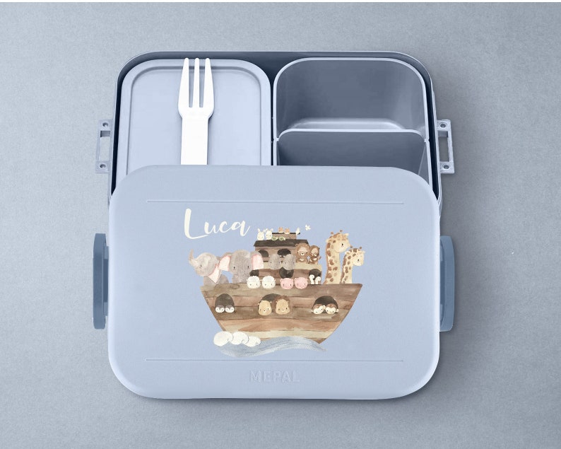 Puede incluir: Una fiambrera azul con tapa extra&iacute;ble y un compartimento separado para cubiertos. La fiambrera tiene una ilustraci&oacute;n colorida de animales en un barco con el nombre "Luca" impreso en la parte delantera.
