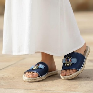 May include: Dark blue denim slide sandals featuring a decorative bee embellishment on the upper strap. The bee design includes light blue sequins for wings, a central dark blue stone, and small metallic accents. The soles have a woven jute trim.