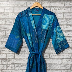 Könnte beinhalten: Ein blauer Patchwork-Bademantel mit passendem Gürtel hängt an einem Holzbügel vor einer weißen Backsteinmauer. Der Bademantel weist verschiedene Blautöne mit dekorativen Nähten und gemusterten Flicken auf. Der Bademantel hat weite Ärmel und einen V-Ausschnitt.