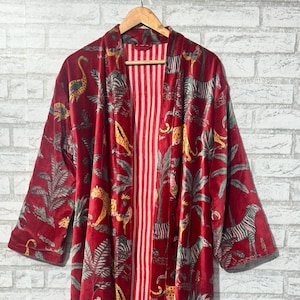 May include: A long, red velvet robe with a jungle animal print, including leopards and zebras, and a striped red and white interior. The robe is hanging on a wooden hanger against a white brick wall.
