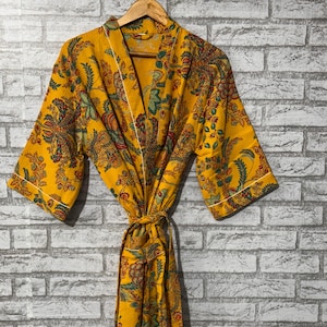 Op de afbeelding: Een gele kimono met een bloemenpatroon in groen, rood en blauw. De kimono heeft een witte bies en een bijpassende strik. De kimono hangt aan een houten hanger tegen een grijze bakstenen muur.