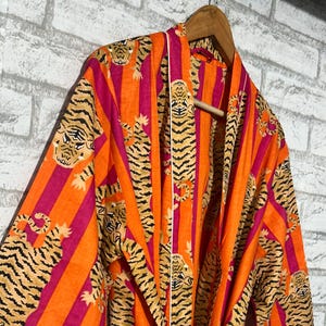 May include: An orange and fuchsia striped robe with a tiger print. The robe is hanging on a wooden hanger against a white brick wall. The robe has white trim and a matching belt.