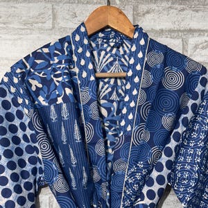 May include: A blue and white patterned robe with a wooden hanger. The robe features various designs, including polka dots, leaf patterns, and geometric shapes. The robe is tied at the waist with a matching fabric belt.