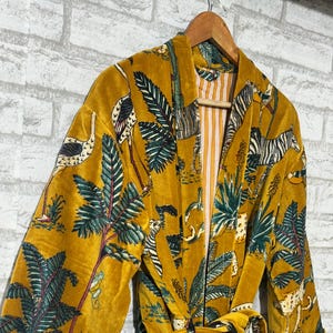 Op de afbeelding: Mosterdgele fluwelen badjas met een jungle dierenprint met zebra's, luipaarden en vogels. De badjas heeft een bijpassende riem en gestreepte manchetten. De badjas hangt aan een houten hanger.
