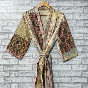 May include: A beige, patterned kimono robe with a tie waist, hanging on a wooden hanger. The robe features a patchwork design with geometric and floral patterns in shades of brown, red, and green. The robe is made of a quilted fabric.