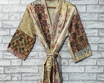 Puede incluir: Bata kimono beige estampada con cinturón anudado, colgada de una percha de madera. La bata presenta un diseño de patchwork con patrones geométricos y florales en tonos marrón, rojo y verde. La bata está hecha de tela acolchada.