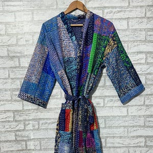 May include: A long-sleeved robe featuring a patchwork design composed of various blue, green, red, and purple fabric squares with intricate patterns. The garment includes a matching tie belt at the waist and visible white running stitch details.