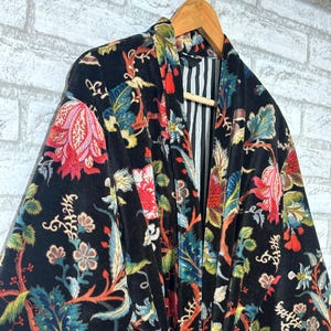 May include: Black velvet robe with a colorful floral pattern in red, blue, and green. The robe has a striped lining and a matching belt with a decorative buckle. The robe is hanging on a wooden hanger.