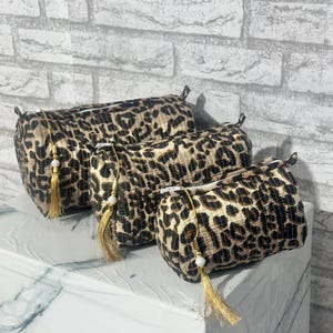 May include: Three leopard print cosmetic bags of varying sizes. Each bag has a gold zipper pull with a gold tassel and a white bead. The bags are cylindrical and sit on a white and gray surface.