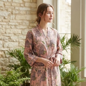 May include: A knee-length robe in a soft pink hue featuring a repeating pattern of dark gray leopards and green palm leaves. The garment includes long sleeves, a V-neckline with white piping, two front pockets, and a matching fabric tie belt.