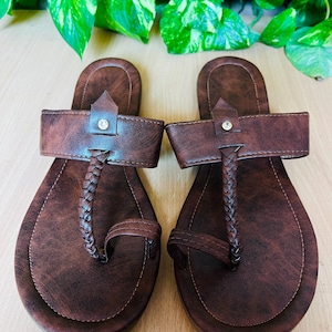 May include: A pair of dark brown flat sandals featuring a faux leather finish with a marbled texture. Each sandal has a wide horizontal strap with a central braided vertical strap, a toe loop, and a small silver-toned circular stud detail.