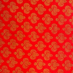 May include: A vibrant red fabric featuring a repeating pattern of golden-orange stylised floral motifs. The design consists of symmetrical, rounded shapes arranged in a grid, creating a textured appearance across the entire surface of the textile.