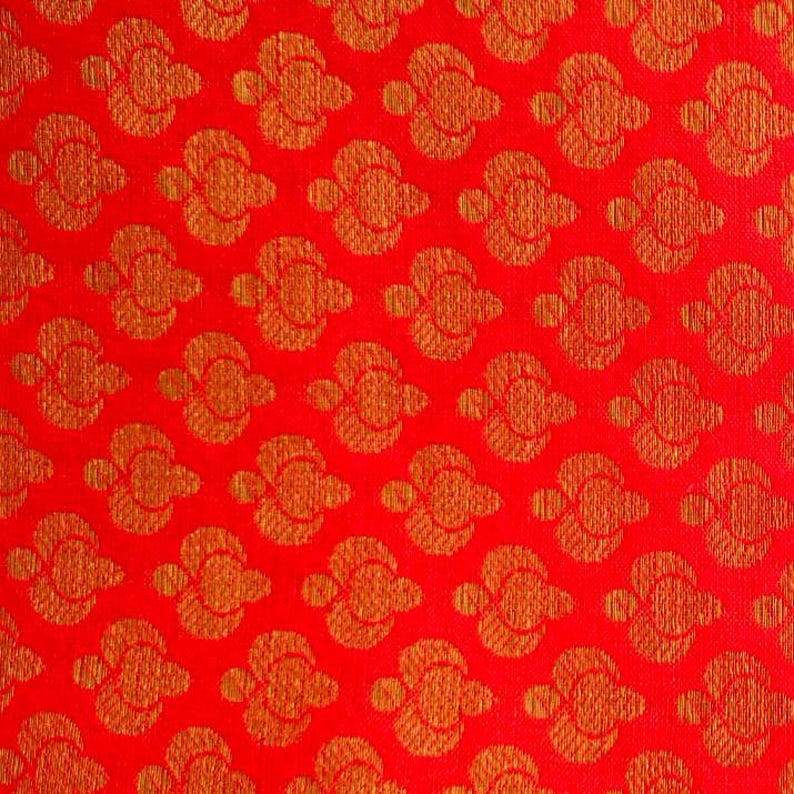 May include: A vibrant red fabric featuring a repeating pattern of golden-orange stylised floral motifs. The design consists of symmetrical, rounded shapes arranged in a grid, creating a textured appearance across the entire surface of the textile.
